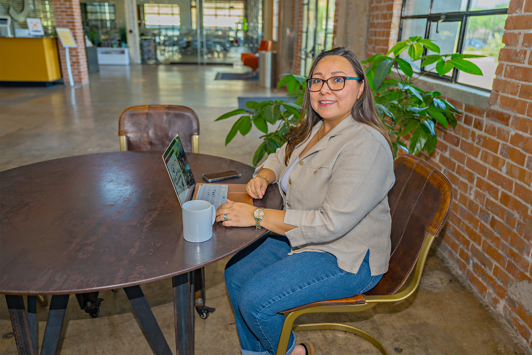 A woman sitting in front of a laptop with cup and a cell phone surrounding it and she's looking at camera